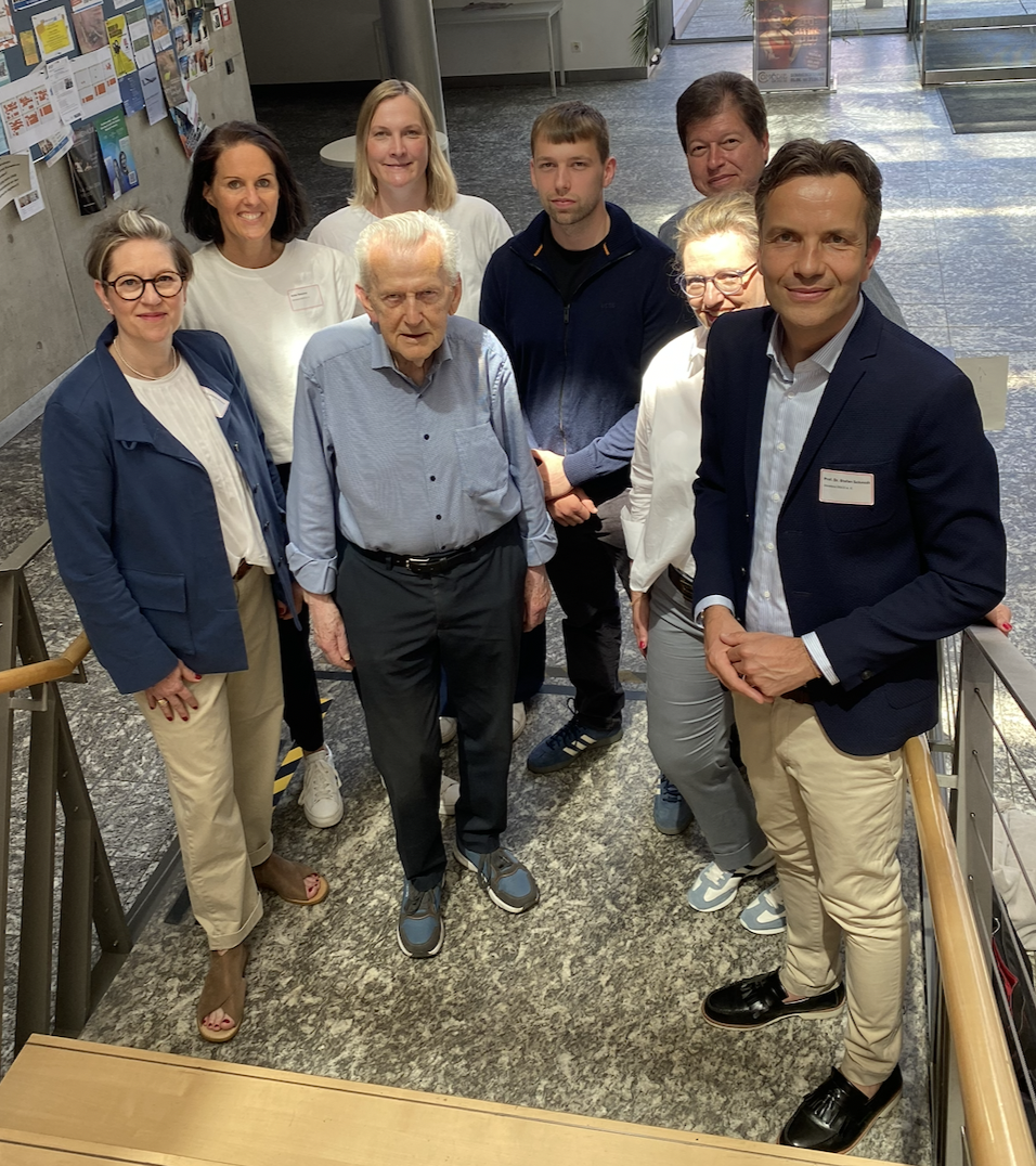 Acht Personen lächeln freundlich in die Kamera. Es handelt sich um eine leichte Draufsicht. Die Kamera wurde auf einer Treppe platziert, die im Foto durch ihr Gelände an den rechten und linken Bildrändern sichtbar wird.