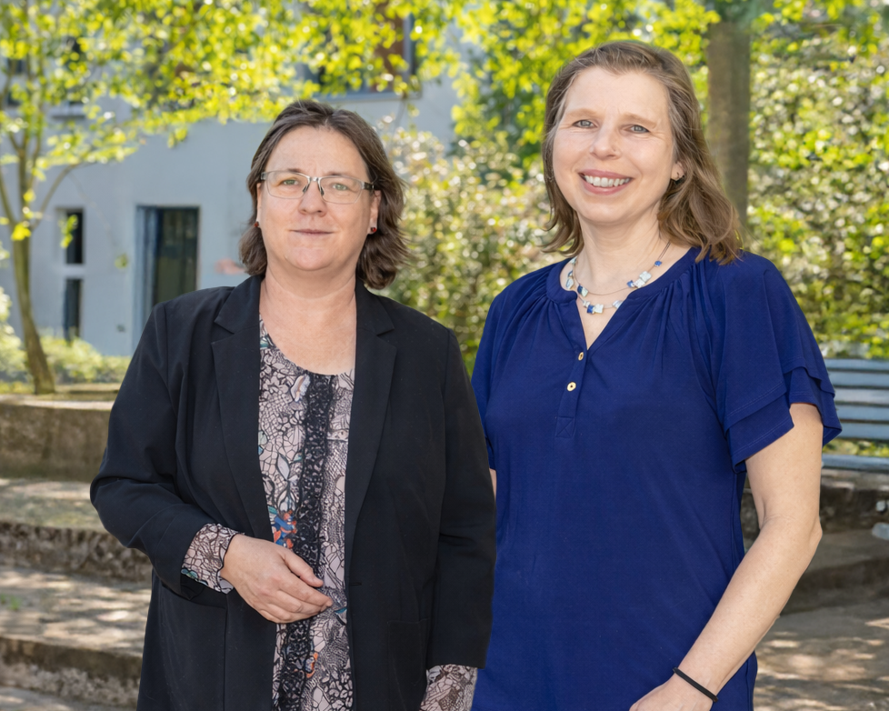 Zwei Frauen lächeln freundlich in die Kamera. Sie stehen in einem Hof mit grünen Bäumen und grauen Betonplatten. Im Hintergrund sind eine weiße Hausfassade und eine blaue Parkbank zu erkennen.