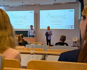 Zwei formell gekleidete Personen stehen in einem Hörsaal. Sie sprechen zu Gästen, die auf den Hörsaalstühlen Platz genommen haben. Hinter der Rednerin und dem Redner sieht man eine Präsentation an der Wand mit dem Titel „Welcome“.