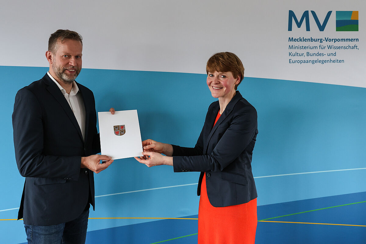 Das Foto zeigt, wie Staatssekretärin Susanne Bowen einen neuen Professoren für die Hochschule Neubrandenburg ernennt. Beide Personen schauen in die Kamera. Eine Urkunde wird übergeben.