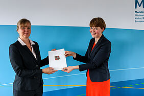 Das Foto zeigt, wie Staatssekretärin Susanne Bowen eine neue Professorin für die Hochschule Neubrandenburg ernennt. Beide Personen schauen in die Kamera. Eine Urkunde wird übergeben.