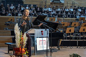 Wissenschaftsministerin Bettina Martin spricht an einem Pult mit der Aufschrift "Hochschule Neubrandenburg". Im Hintergrund befinden sich ein großes Piano und ein Chor.
