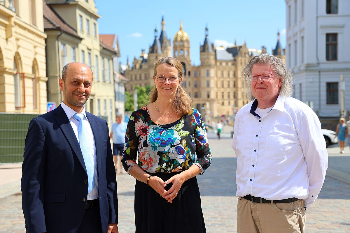 Ministerin Bettina Martin steht, zusammen mit den frisch ernannten Professoren zu einem Gruppenfoto zusammen. Im Hintergrund strahlt das Schweriner Schloss in sommerlicher Sonne und hellblauem Himmel.
