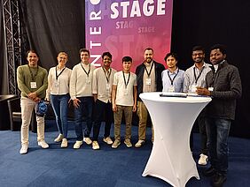 Auf dem Gruppenfoto befinden sich neun Personen, die lächelnd in die Kamera schauen. Alle Personen tragen Lanyards mit Messeausweisen um den Hals.