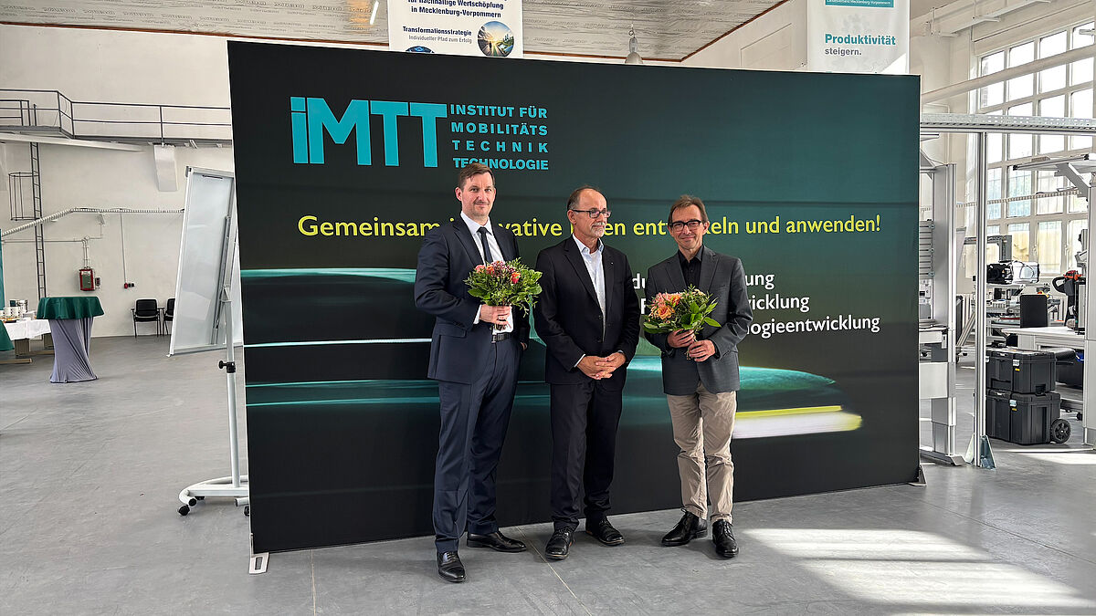 Drei Personen stehen vor einer dunklen Fotowand mit der Aufschrift "Institut für Mobilitätstechnik". Zwei der drei sehr förmlich gekleideten Männer halten je einen Blumenstrauß in der Hand und lächeln freundlich in die Kamera.