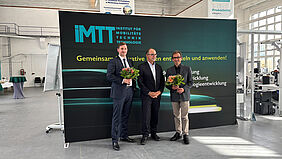 Drei Personen stehen vor einer dunklen Fotowand mit der Aufschrift "Institut für Mobilitätstechnik". Zwei der drei sehr förmlich gekleideten Männer halten je einen Blumenstrauß in der Hand und lächeln freundlich in die Kamera.