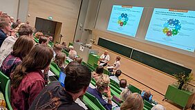 Zwei Frauen sitzen in einem gut gefüllten Hörsaal auf Hockern neben einem Rednerpult. Eine der Personen hält ein Mikrofon in der Hand. Im Hintergrund wir eine Präsentation an die Wand projiziert, die sich mit der Bewirtschaftung oberirdischer Gewässer befasst.