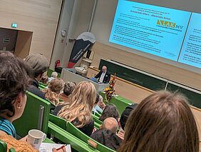 Zu sehen ist ein Mann in weißem Hemd und dunklem Sakko, der an einem Pult vor einer grünen Tafel steht. Über der Tafel wird eine Präsentation mit dem Titel „Armutsrisiko Altern – Altersrisiko Armut“ abgespielt. Die grünen Bänke im Hörsaal sind vor dem Referenten gut gefüllt. 