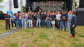 Zu sehen sind Menschen, die vor einer Bühne stehen und ihren Sieg feiern. Einer der Personen hält einen Pokal, eine andere hält eine Urkunde in die Kamera.