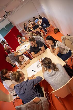 Das Foto zeigt einen Seminarraum mit größeren Tischen, an denen jeweils fünf bis sechs Personen sitzen. Die Personen sind locker gekleidet, lächeln einander an. 