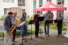 echs Saxophonisten spielen an offener Luft. Vor ihnen stehen verschieden hohe Notenständer. Im Hintergrund steht ein rotes Pavillon mit der Aufschrift „Fachschaftsrat Gesundheit, Pflege, Management“.