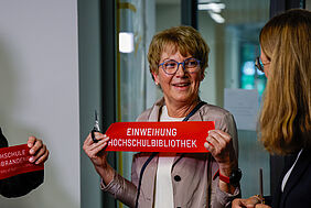 Eine Frau hält ein rotes Band mit der Aufschrift "Einweihung Hochschulbibliothek" lächelt freudestrahlend eine weitere Person an. Die Frau trägt kurzes blondes Haar, eine blaue, runde Brille und einen hellen Blazer über weißem Oberteil.