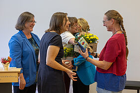 Fünf Frauen stehen sich gegenüber. Die Situation ist freundlich und wohlwollend. Im Hintergrund findet eine Umarmung statt und es werden Blumensträuße überreicht.