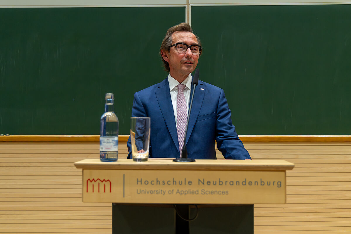 Ein Mann, der einen blauen Anzug und ein weißes Hemd trägt, steht vor einem Pult, auf dem eine Wasserflasche und ein leeres Glas steht. Hinter ihm befindet sich eine grüne Kreidetafel.
