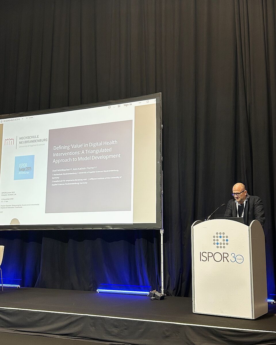 Ein Mann steht hinter einem Pult mit der Aufschrift „ISPOR 30“. Hinter ihm hängt ein schwerer, schwarzer Vorhang in Falten. Neben ihm befindet sich ein großer Bildschirm auf der Bühne. Dieser zeigt eine Präsentation mit dem Logo der Hochschule Neubrandenburg.