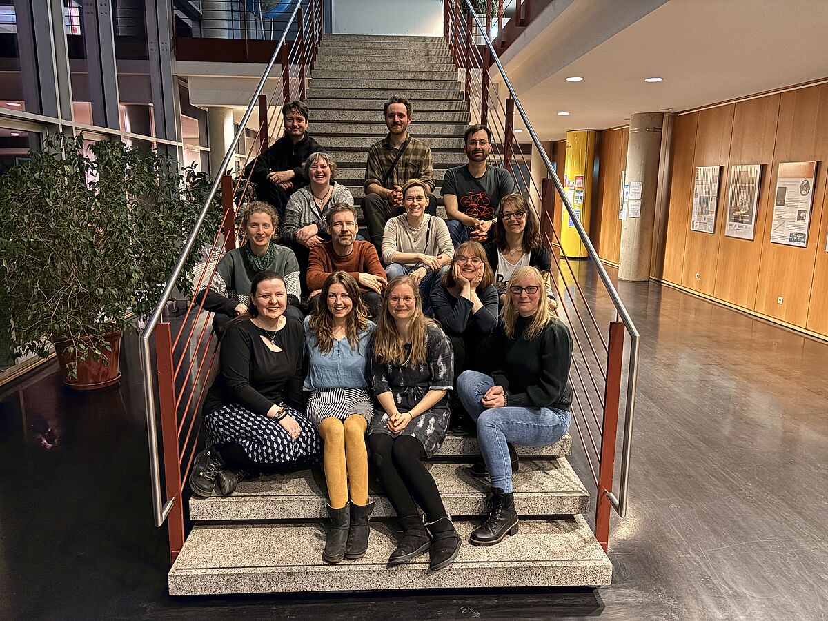 Auf dem Foto befinden sich 13 Personen, auf einer Treppe sitzend. Es handelt sich um ein lockeres Gruppenfoto. Die Personen lächeln in die Kamera.
