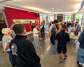 Ein Raum, der auf mindestens einer Seite durch Tageslicht gespeist wird, ist gefüllt mit Menschen. Die vorrangig Jugendlichen reden und stehen an einem der aufgebauten Stände an.