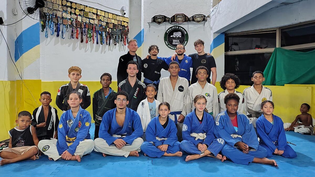 Auf diesem Gruppenfoto sind Trainer, Kinder und Jugendliche eines Jiu-Jitsu-Kurses zu sehen. Die Personen haben sich in drei Reihen positioniert und tragen klassische Kampfsportroben in schwarz, weiß und blau. Das Foto wurde wahrscheinlich in einer Sport- oder Trainingsstätte aufgenommen. An den Wänden befinden sich Auszeichnungen wie Medaillen und Meistergürtel.