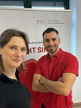 Juliane Häntze, Studiengangskoordinatorin Bauingenieurwesen und Tom Gehlhar, Studiengangleiter Bauingenieurwesen lächeln in die Kamera. Im Hintergrund ist ein weiß-roter Rollup der Hochschule zu sehen.