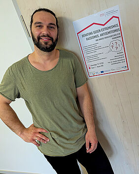 Ein Mann steht lächelnd vor einer Tür. Er trägt eine schwarze enge Jeans und ein weit geschnittenes grünes Shirt. An der Tür hängt ein Poster, das auf ein Beratungsangebot an der Hochschule aufmerksam macht.