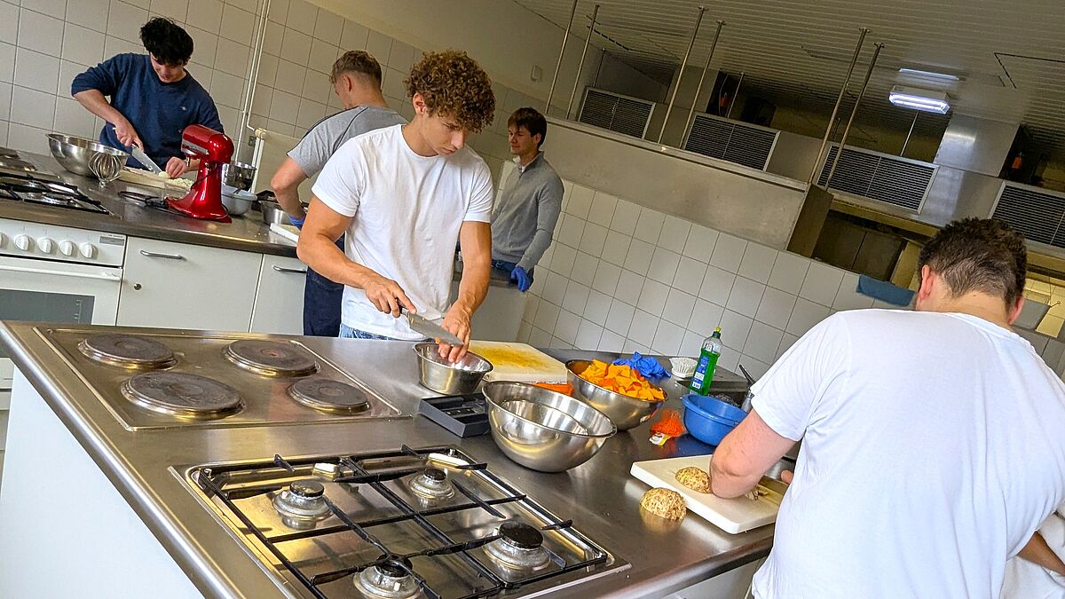 Das Foto zeigt mehrere Personen, vorrangig Jugendliche, die an verschiedenen Kochinseln arbeiten. Die Kochinseln haben metallische Oberflächen wie in einer Großküche. Auf den Oberflächen befinden sich Schüsseln, Rührgeräte, Schneidebretter und Messer.