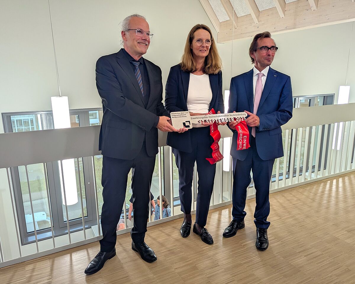 Drei Personen stehen nebeneinander und lächeln den vermutlich anwesenden Fotografen zu. Ministerin Bettina Martin steht in der Mitte und überreicht symbolisch einen großen silbernen Schlüssel als Zeichen der Übergabe der Hochschulbibliothek Neubrandenburg.