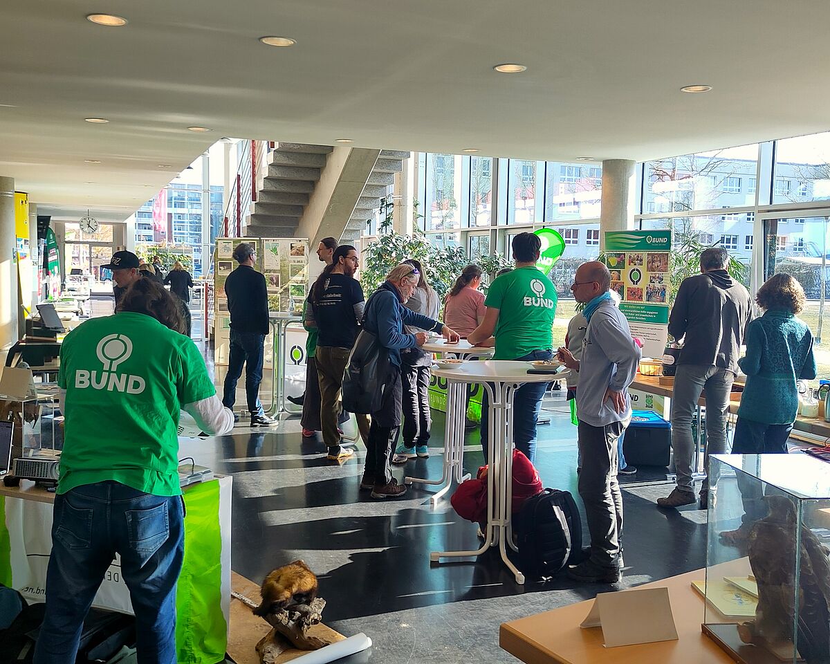 Zu sehen ist das lichtdurchflutete Foyer vom Haus 2 der Hochschule. Mindestens 15 Personen stehen an verschiedenen Punkten im Raum. Verschiedene Banner, Aufsteller  und Infotafeln lassen darauf schließen, dass es sich um eine messeähnliche Veranstaltung handelt.