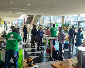 Zu sehen ist das lichtdurchflutete Foyer vom Haus 2 der Hochschule. Mindestens 15 Personen stehen an verschiedenen Punkten im Raum. Verschiedene Banner, Aufsteller  und Infotafeln lassen darauf schließen, dass es sich um eine messeähnliche Veranstaltung handelt.