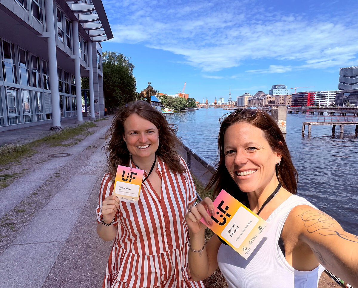 Zwei lächelnde Frauen stehen am Ufer der Spree in Berlin und halten ihre farbigen Namensschilder in die Kamera. Die Frau rechts macht ein Selfie und trägt ein weißes ärmelloses Oberteil, die Frau links ein rot-weiß gestreiftes Kleid. Im Hintergrund sind das Wasser, moderne Gebäude und der Berliner Fernsehturm unter einem sonnigen Himmel mit vielen Wolken zu sehen.