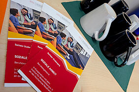 Zu seheb sind aufgefächerte Flyer zum berufsbegleitenden Studiengang Soziale Arbeit. Im Hintergrund befinden sich schwarze und weiße Tassen, die darauf schließen lassen, dass es vor Ort auch Kaffee gibt.
