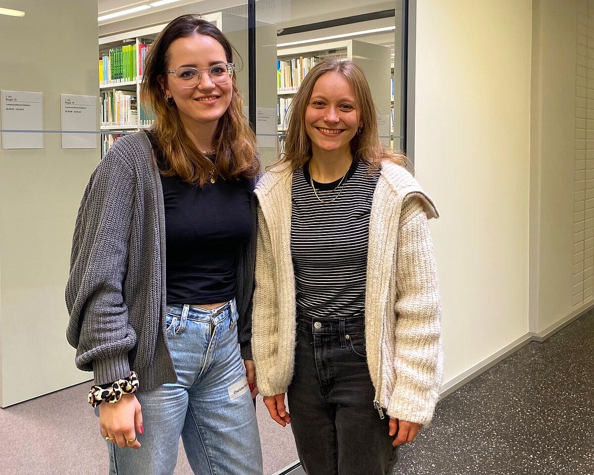 Zwei leger gekleidete Frauen lächeln freundlich in die Kamera. Im Hintergrund ist ein Fenster zur Hochschulbibliothek zu erkennen.