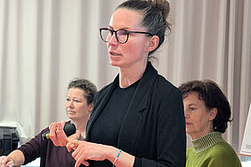 Eine Frau spricht zu einem Publikum, welches auf dem Foto nicht zu sehen ist. Hinter ihr sitzen zwei weitere Frauen mit Blick Richtung Publikum.