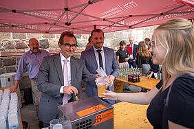 Der Rektor der Hochschule und der Oberbürgermeister Neubrandenburgs stehen unter einem roten Pavillon und schenken Bier aus. Im Hintergrund sind weitere Getränke in Flaschen abgefüllt. Im Vordergrund steht eine junge Frau, die dem Rektor einen gefüllten Becher abnimmt. Sie lächelt. 