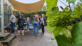 Mehrere Menschen stehen und sitzen gemeinsam und miteinander im Gespräch unter einem Sonnensegel. Im Hintergrund befindet sich eine große grüne Hecke, im Vordergrund ist unscharf eine Weinrebe zu erkennen. Am linken Bildrand ist ein Gewächshaus zu erkennen, dessen Tür geöffnet ist.