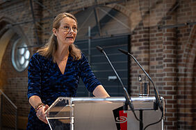 Ministerin Bettina Martin steht an einem Redepult und spricht zum Publikum. Hinter ihr sind die schweren Backsteinmauern der Konzertkirche Neubrandenburg zu erkennen.