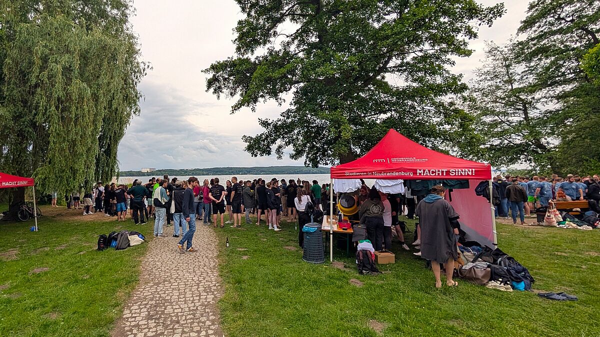 Zu sehen ist eine Außenaufnahme von einem Strandabschnitt voller Menschen. Ein roter Pavillon mit Der Aufschrift „Studieren in Neubrandenburg“ steht im Vordergrund. Im Hintergrund ist der Tollensesee zu erkennen.