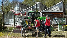 Ein sommerlicher Tag. Im Hintergrund befindet sich ein großes Glashaus, im Vordergrund stehen Landmaschinen und Menschen in roten Westen. 