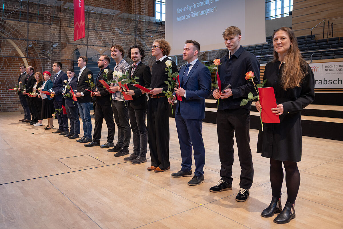 Feierlich gekleidete Personen stehen aufgereiht nebeneinander auf einer Bühne. In den Händen halten sie ihr Abschlusszeugnis, die Ehrenmünze sowie eine Rose. 