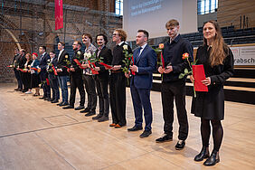 Feierlich gekleidete Personen stehen aufgereiht nebeneinander auf einer Bühne. In den Händen halten sie ihr Abschlusszeugnis, die Ehrenmünze sowie eine Rose. 