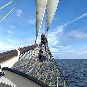 Das Foto zeigt drei Personen, die am vorderen Ende eines Segelschiffs herum klettern. Unter ihnen befindet sich ein Netz aus dicken Seilen und das wellige Meer. Der Himmel ist leicht bewölkt und satt blau.