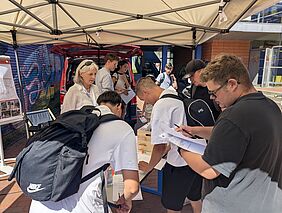 Mehrere Schülerinnen, Schüler und Auszubildende stehen unter einem Pavillon an einem Informationsstand, bearbeiten ein Quiz, beschäftigen sich mit Informationsmaterial. Mitarbeitende des Studiengangs beraten die Teilnehmenden und beantworten Fragen zum Studium der Geodäsie und Messtechnik.