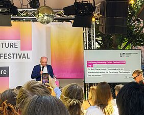Ein Mann im blauen Anzug steht auf einer Bühne und spricht zum Publikum. Hinter ihm hängen große Banner in den Farben Pink, Orange und Gelb des University:Future Festivals. Rechts zeigt ein Bildschirm den Programmpunkt „Eröffnung University:Future Festival 2026 – Tag 2“ mit dem Namen des Redners. Im Vordergrund sitzen zahlreiche Besucherinnen und Besucher. Über der Bühne hängen Scheinwerfer und eine Discokugel.
