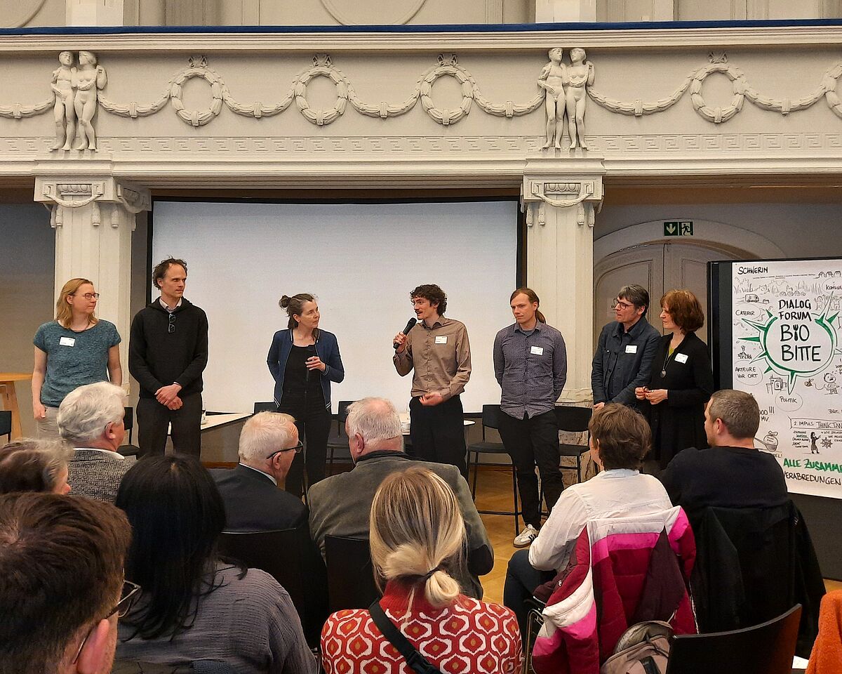 Mehrere Personen stehen vor sitzendem Publikum in einem opulenten, hellen Saal. Ein Mann hält ein Mikrofon und spricht direkt zu den Anwesenden.