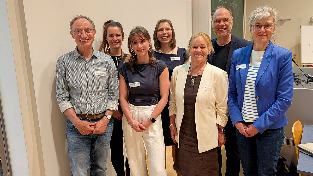 Ein Gruppenfoto. Sieben Personen stehen, aufgereiht in zwei Reihen, dicht nebeneinander und lächeln freundlich in die Kamera. Alle sind eher förmlich gekleidet. Im Hintergrund ist eine klassische, grüne Schultafel zu erkennen.