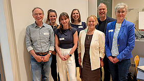 Ein Gruppenfoto. Sieben Personen stehen, aufgereiht in zwei Reihen, dicht nebeneinander und lächeln freundlich in die Kamera. Alle sind eher förmlich gekleidet. Im Hintergrund ist eine klassische, grüne Schultafel zu erkennen.