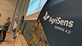 Zu sehen ist Moritz von Oertzen, Landwirt und Referent des Ergebnis-Symposiums des Projekts AgriSens-DEMMIN 4.0. Er steht vor einer Tafel im Hörsaal. Im Vordergrund befindet sich eine Flagge des Digitalisierungsprojekts.