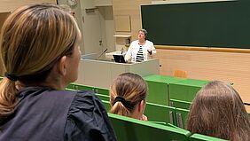 Eine Frau steht hinter einem Rednerpult in einem Hörsaal. Sie trägt eine helle Jacke und ein gestreiftes Oberteil. Hinter ihr befindet sich eine große grüne Tafel. Die Personen im Publikum sind nur von hinten erkennbar. Ganz links im Bildrand ist eine Frau mit bräunlichen Haaren und einem Zopf zu erkennen.