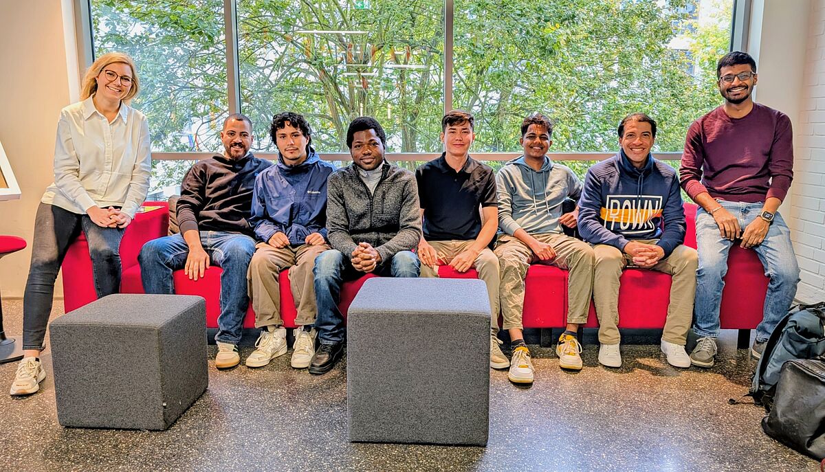 Acht Personen sitzen auf einem roten Sofa vor einer Fensterfront. Im Hintergrund befinden sich grüne Bäume. Die Personen lächeln freundlich in die Kamera.