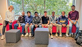 Acht Personen sitzen auf einem roten Sofa vor einer Fensterfront. Im Hintergrund befinden sich grüne Bäume. Die Personen lächeln freundlich in die Kamera.