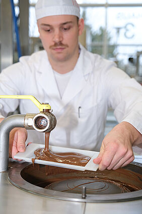 Zu sehen ist ein junger Lebensmitteltechnologe in weißerArbeitskleidung an einer Maschine, aus der flüssige Schokolade fließt. Mit einer weißen Tafelform fängt er die Schokolade auf. Im Hintergrund befinden sich helle, große Fenster.